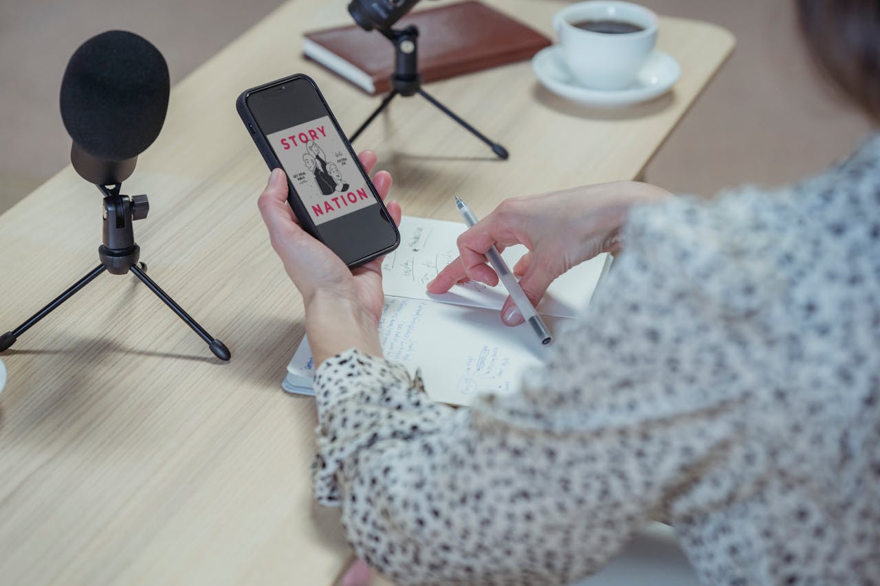 our-story A woman records a podcast at a desk using a smartphone and microphones.